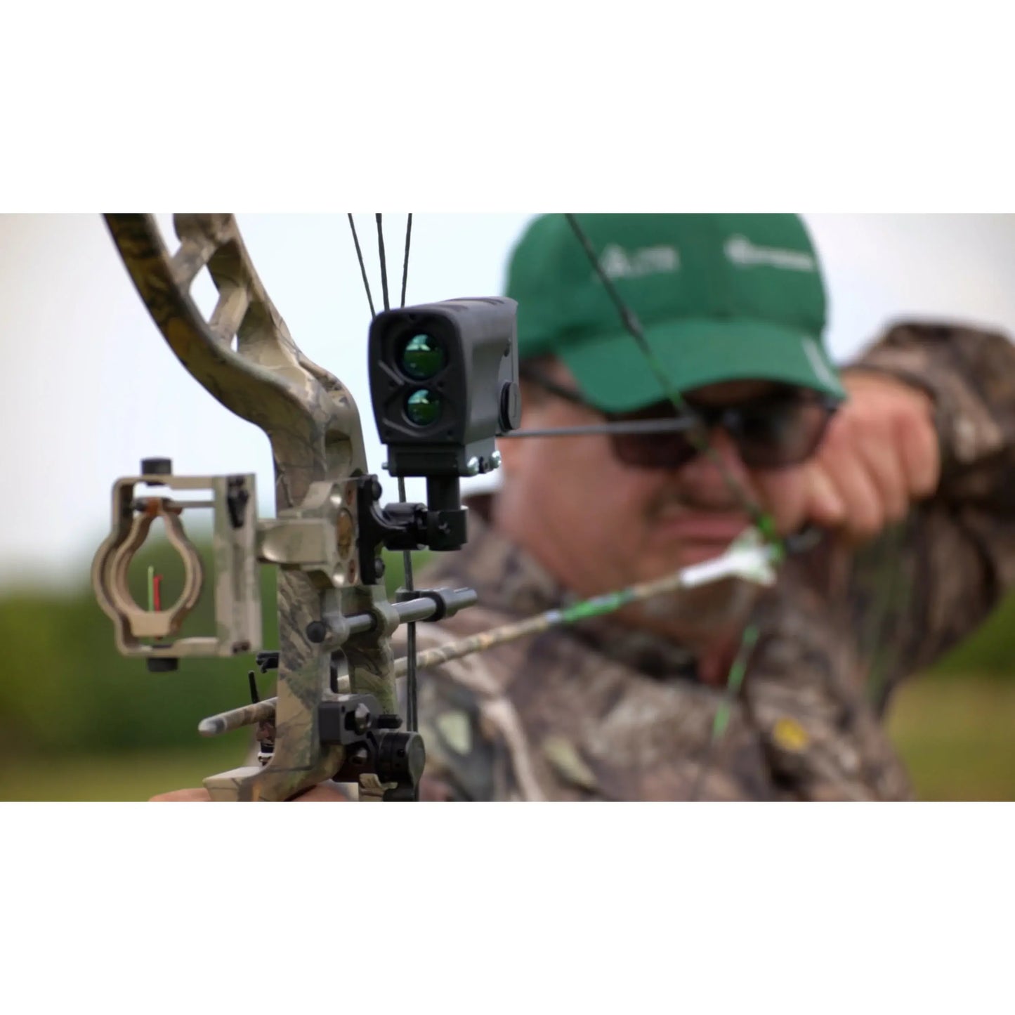 Person in camouflage gear using a bow with a sight, wearing a green cap.