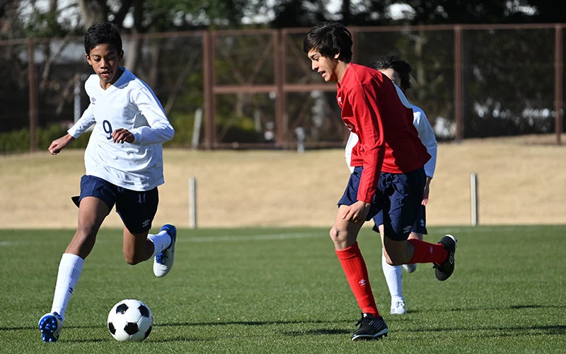 Two young soccer players in action on a grass field with a fence in the background.