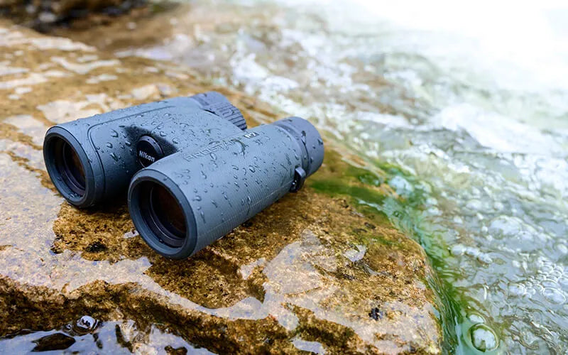 Nikon Prostaff P7 binoculars on a wet rock covered in water droplets with moving water in the background.