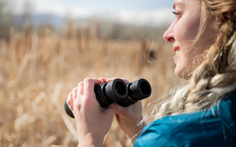 Person using binoculars in a natural setting with tall grass and blurred background