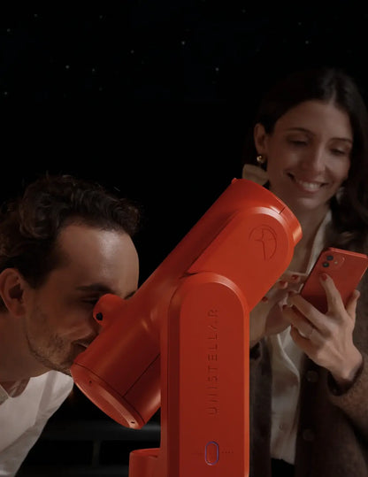Two people using a red telescope and phone against a dark background
