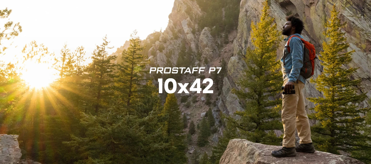 Person standing on a rocky outcrop with mountains and trees in the background, wearing a Prostaff P7 binoculars package.