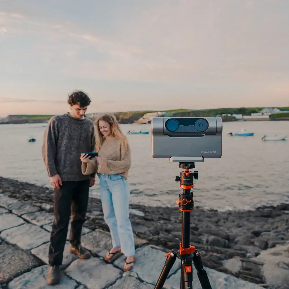 Two people standing on a rocky shore with a smartphone and a DWARF Telescope on a tripod in the foreground.