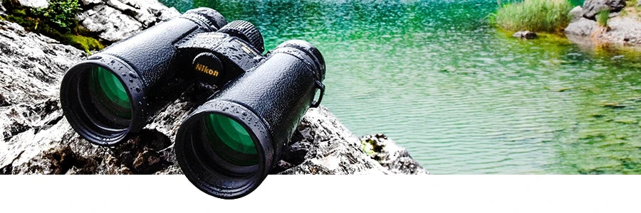 A wet set of MONARCH HG binoculars on some rocks with a stream in the background.