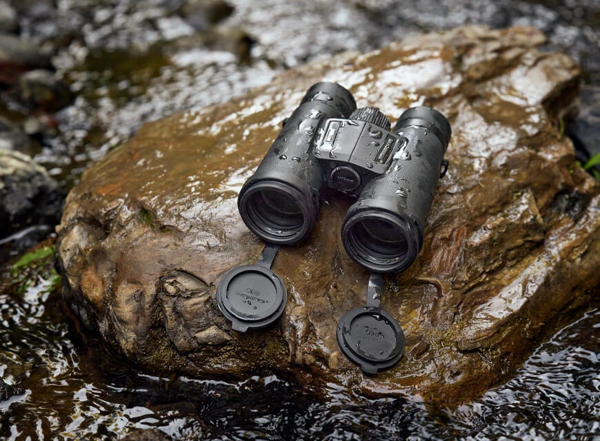 Nikon binoculars are wet and perched on a wet rock with moving water from a creek in the background.